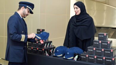 An Emirates pilot in uniform receives a commemorative cap from a staff member during World Pilots' Day celebrations, with branded ceramic mugs and caps displayed on a table.