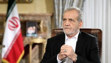 Iranian President Masoud Pezeshkian sits during a formal address, hands clasped, with the Iranian flag and a portrait in the background.