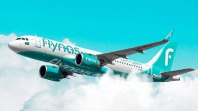 The image of a Flynas airplane, with a white and teal color scheme, flies through a partly cloudy sky.