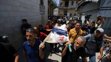 The image of a group of mourners, including men in casual clothing, carry a stretcher with the body of Palestinian journalist Ismail Baddah, draped in a white cloth and a blue jacket, through a narrow street in Gaza City.