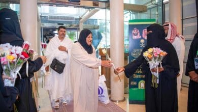 A female pilgrim receives a flower from a Saudi official as she arrives for Haj, with other pilgrims following behind in a warmly decorated arrival area.