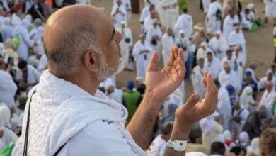 A Haj pilgrim in white ihram raises his hands in prayer, surrounded by a large crowd of fellow worshippers at Mount Arafat.